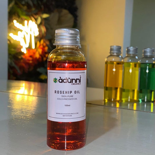 A bottle of Adunni Organics pure cold pressed Rosehip Oil with a label showing the product name and volume, placed on a table with other colored oils in the background.